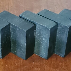 May include: Five dark gray rectangular bars of soap are arranged in a row. The soap bars have a slightly textured surface with small white specks. The edges are clean and sharp, and the overall appearance is simple and modern.