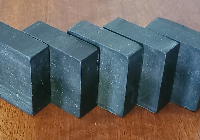 May include: Five dark gray rectangular bars of soap are arranged in a row. The soap bars have a slightly textured surface with small white specks. The edges are clean and sharp, and the overall appearance is simple and modern.