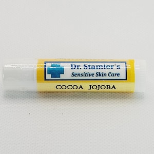 May include: A lip balm tube with a yellow and white label. The label reads "Dr. Stamier's Sensitive Skin Care" with the words "COCOA JOJOBA" below. The lip balm is in a clear plastic tube.