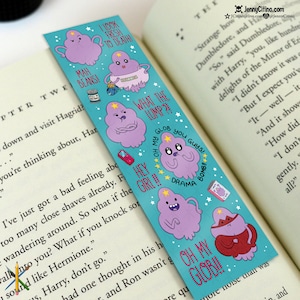 Könnte beinhalten: Ein türkisfarbenes Lesezeichen mit Cartoon-Charakteren mit lila wolkenartigen Körpern und sternförmigen Kronen. Auf dem Lesezeichen stehen Sätze wie "MAH BEANS!" und "OH MY GLOB!!". Das Lesezeichen liegt auf einem offenen Buch.