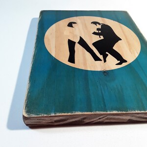 May include: A wooden sign with a teal blue background and a white circle in the center. The circle has two black silhouettes of people dancing in the center.