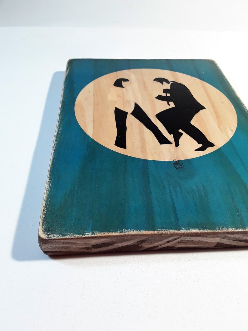 May include: A wooden sign with a teal blue background and a white circle in the center. The circle has two black silhouettes of people dancing in the center.
