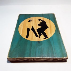 May include: A wooden sign with a teal background and a yellow circle in the center. The circle contains a black silhouette of two people dancing. The sign has a distressed look and is made of wood.