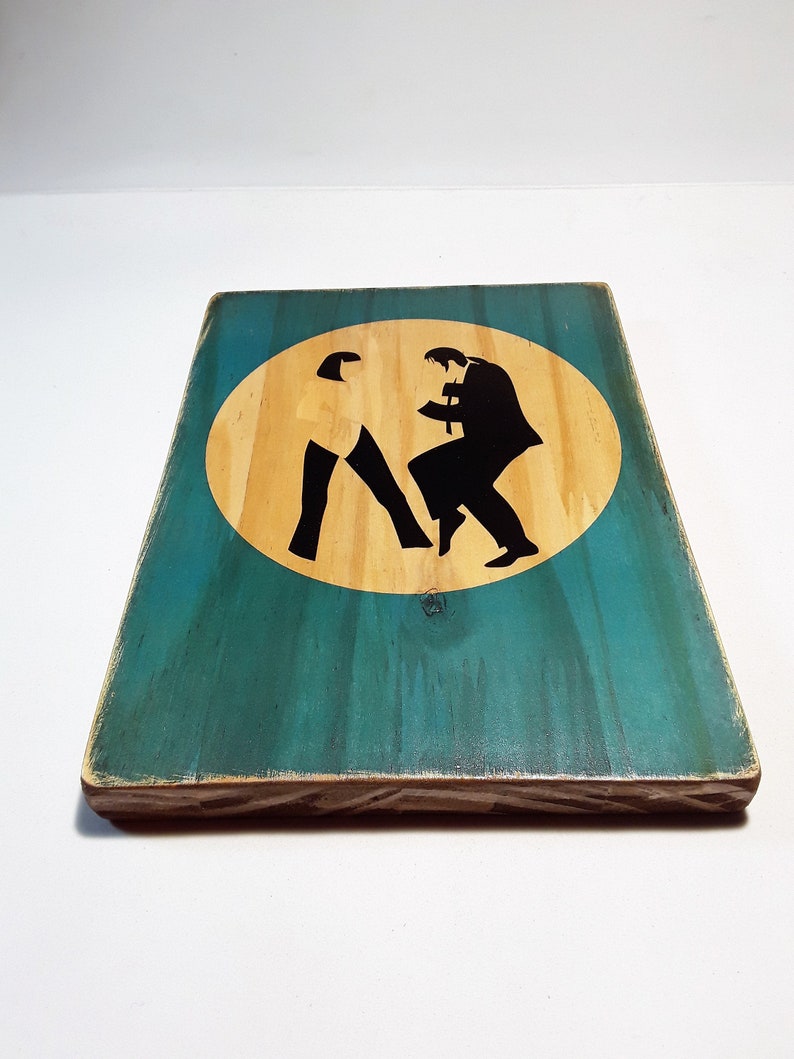 May include: A wooden sign with a teal background and a yellow circle in the center. The circle contains a black silhouette of two people dancing. The sign has a distressed look and is made of wood.