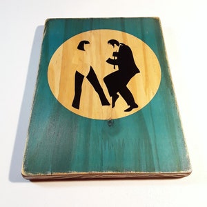 May include: A wooden sign with a teal green background and a circular cutout in the center. The cutout is filled with a light brown wood color and features a black silhouette of two people dancing.