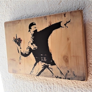Può includere: Una stampa d'arte a stencil in bianco e nero su un pannello di legno che raffigura una persona che lancia un mazzo di fiori. La persona indossa una giacca nera e un berretto nero. Lo sfondo è una venatura di legno chiaro.