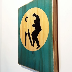 May include: A wooden sign with a teal background and a circular cutout in the center. The cutout is filled with a light wood color and features a silhouette of two people dancing. The sign has a distressed look with visible wood grain.