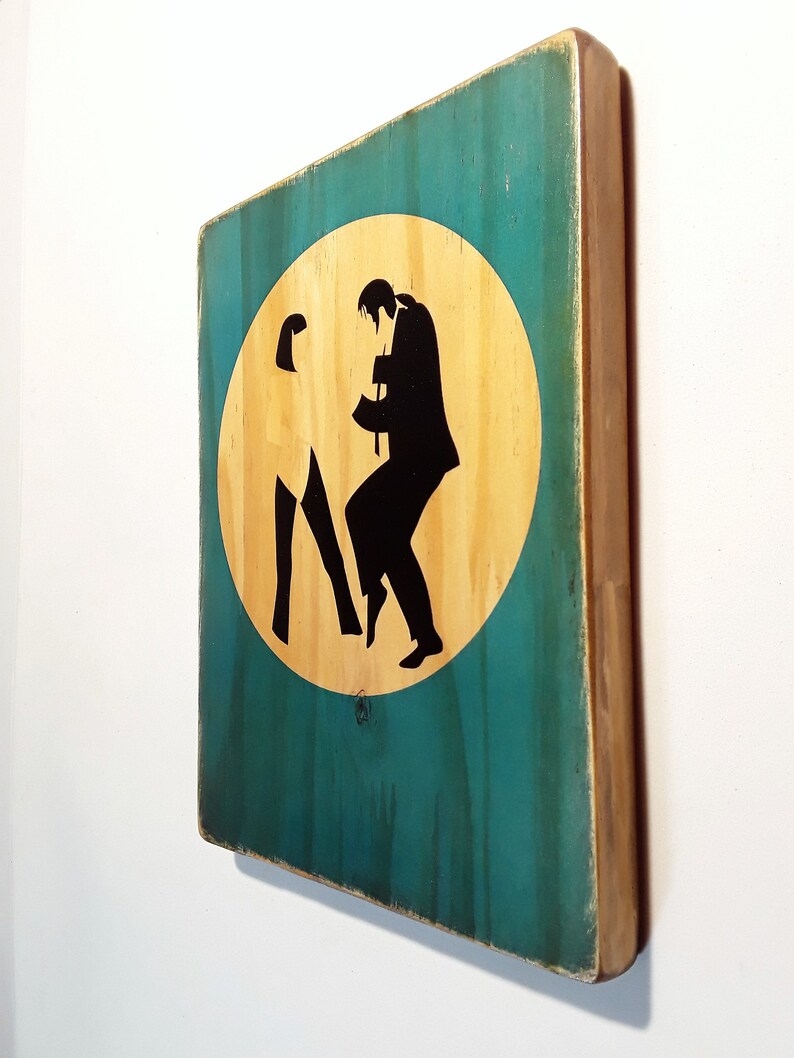 May include: A wooden sign with a teal background and a circular cutout in the center. The cutout is filled with a light wood color and features a silhouette of two people dancing. The sign has a distressed look with visible wood grain.