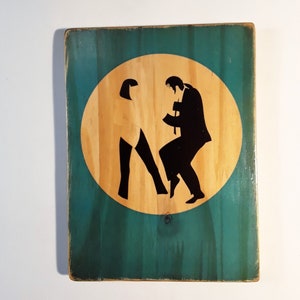 May include: A wooden sign with a teal green background and a circular cutout in the center. The cutout is filled with a light brown wood and features a silhouette of two people dancing. The people are black in color and are positioned in a classic dance pose.