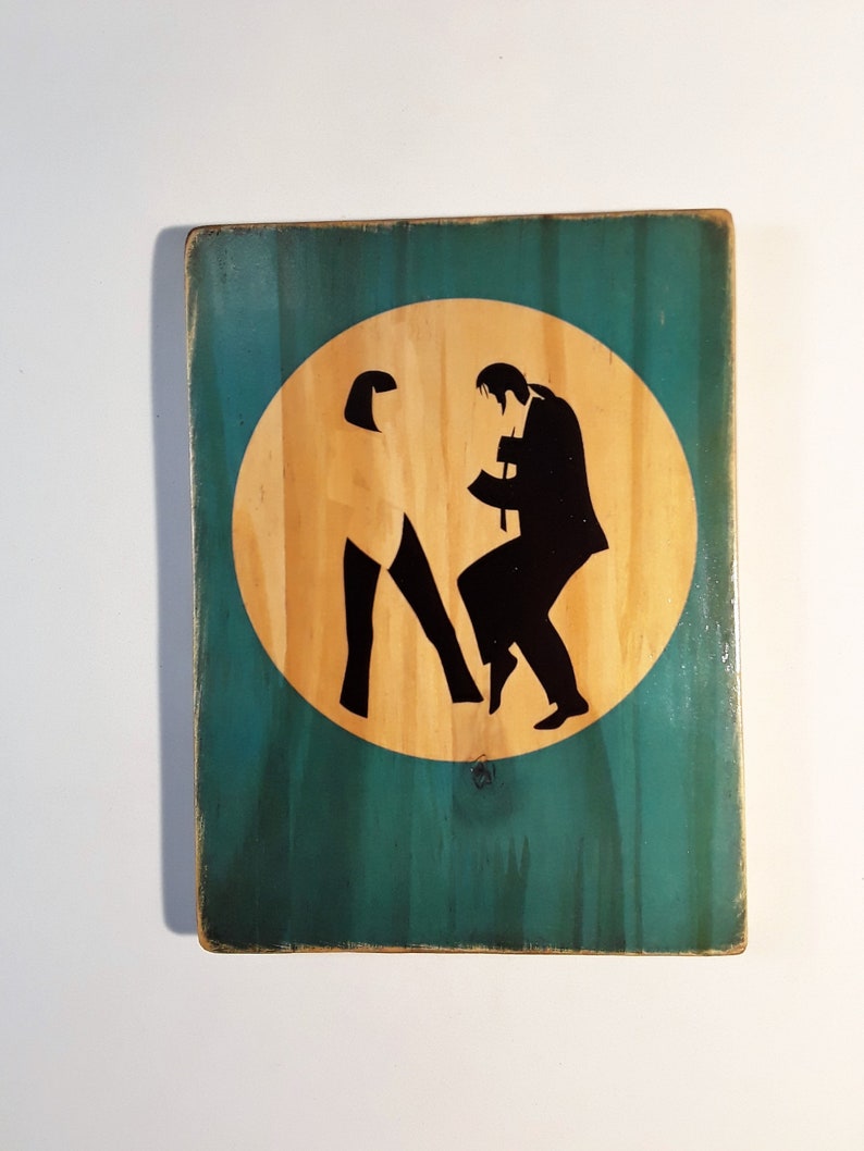 May include: A wooden sign with a teal green background and a circular cutout in the center. The cutout is filled with a light brown wood and features a silhouette of two people dancing. The people are black in color and are positioned in a classic dance pose.