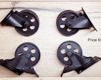 May include: Four black cast iron casters with a vintage industrial design. Each caster has a wheel with five holes and a mounting plate. The casters are arranged on a light brown wooden surface.