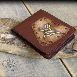 Puede incluir: Cartera de cuero marrón con un parche decorativo en forma de diamante con un diseño de pájaro dorado. El parche está asegurado con pequeñas remaches redondos. La cartera se muestra sobre una superficie de madera.
