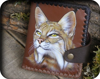 May include: A brown leather wallet featuring a painted image of a bobcat. The bobcat has yellow eyes and a white chin. The wallet has a brown leather strap with a gold metal button.