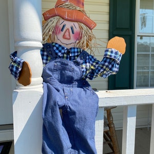 May include: A wooden scarecrow with a straw hat, blue overalls, and a plaid shirt is sitting on a white porch railing. The scarecrow has a painted face with rosy cheeks and a friendly smile.