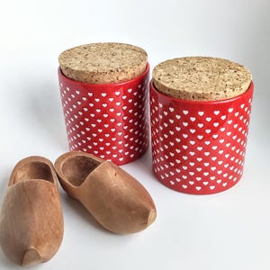 May include: Two red ceramic canisters with white heart patterns and cork lids, next to two wooden clogs.