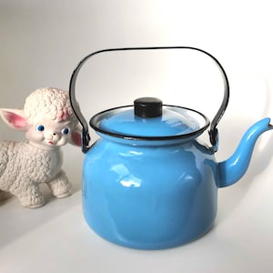 May include: A blue enamel teapot with a black handle and spout. The teapot is sitting on a white surface next to a white rubber toy lamb.
