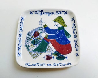 May include: A white ceramic square plate with a blue and white border. The center of the plate features a colorful illustration of a person in a blue jacket and red pants holding a fishing net with fish. The text "Bergen" is written in blue on the plate.