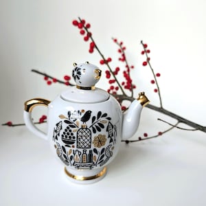 May include: White ceramic teapot with gold detailing on the handle, spout, and base. The teapot has a black and gold floral and bird design. The lid features a black and white floral pattern. Red berries are in the background.