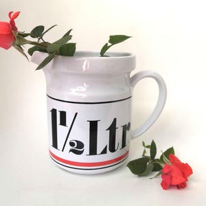 May include: White ceramic jug with a black and red stripe design. The jug has a handle and says "1 1/2 Ltr" on the front. A red rose is partially inside the jug.