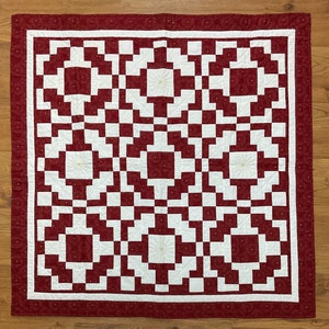 Pode incluir: Um cobertor vermelho e branco com um padrão geométrico. O cobertor é feito de quadrados e retângulos, e o padrão é criado pelas diferentes cores do tecido. O cobertor é enfeitado com uma borda branca.