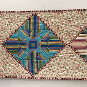 Peut inclure: Un chemin de table matelass&eacute; avec un motif g&eacute;om&eacute;trique dans des tons de rose, bleu, vert et blanc. Le chemin de table est fait de tissu &agrave; imprim&eacute; floral et a une bordure rouge et blanche.