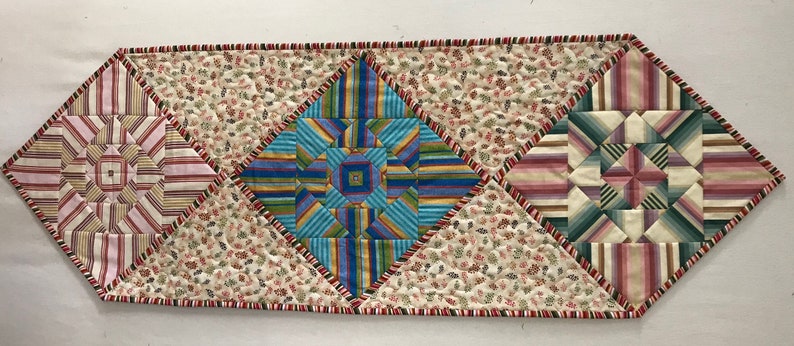 Peut inclure: Un chemin de table matelass&eacute; avec un motif g&eacute;om&eacute;trique dans des tons de rose, bleu, vert et blanc. Le chemin de table est fait de tissu &agrave; imprim&eacute; floral et a une bordure rouge et blanche.