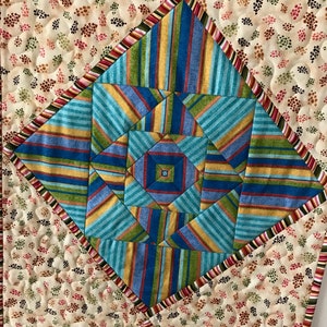 Peut inclure: Un chemin de table matelass&eacute; avec un motif g&eacute;om&eacute;trique dans des tons de bleu, vert, jaune et orange. Le centre du chemin de table pr&eacute;sente un motif en &eacute;toile. Le chemin de table est entour&eacute; d'une bordure de rayures roses, rouges et vertes.