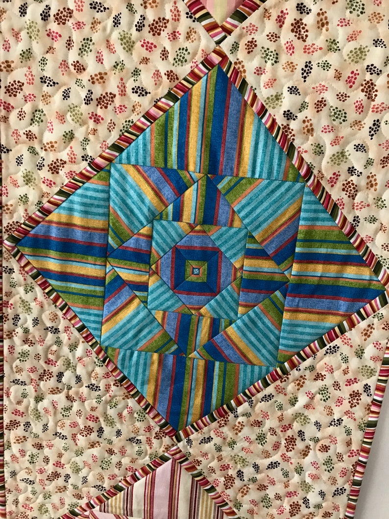 Peut inclure: Un chemin de table matelass&eacute; avec un motif g&eacute;om&eacute;trique dans des tons de bleu, vert, jaune et orange. Le centre du chemin de table pr&eacute;sente un motif en &eacute;toile. Le chemin de table est entour&eacute; d'une bordure de rayures roses, rouges et vertes.