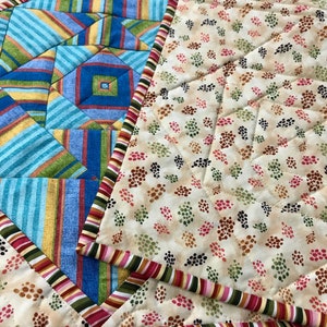 Peut inclure: Un dessous de plat matelass&eacute; avec un motif g&eacute;om&eacute;trique dans des tons de bleu, jaune et vert. Le dessous de plat a une bordure de rayures rouges, roses, vertes et blanches.