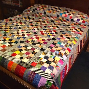 May include: A colorful patchwork quilt with a black and white striped border. The quilt is made of many different fabrics and patterns, and it is laid out on a bed.