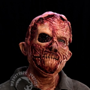 May include: A full-head latex mask featuring a highly detailed, realistic zombie design with exposed muscle tissue, deep red and brown skin tones, and a hollowed-out eye socket. The mask includes a logo reading MOONSTRUCK STUDIOS on the collar.