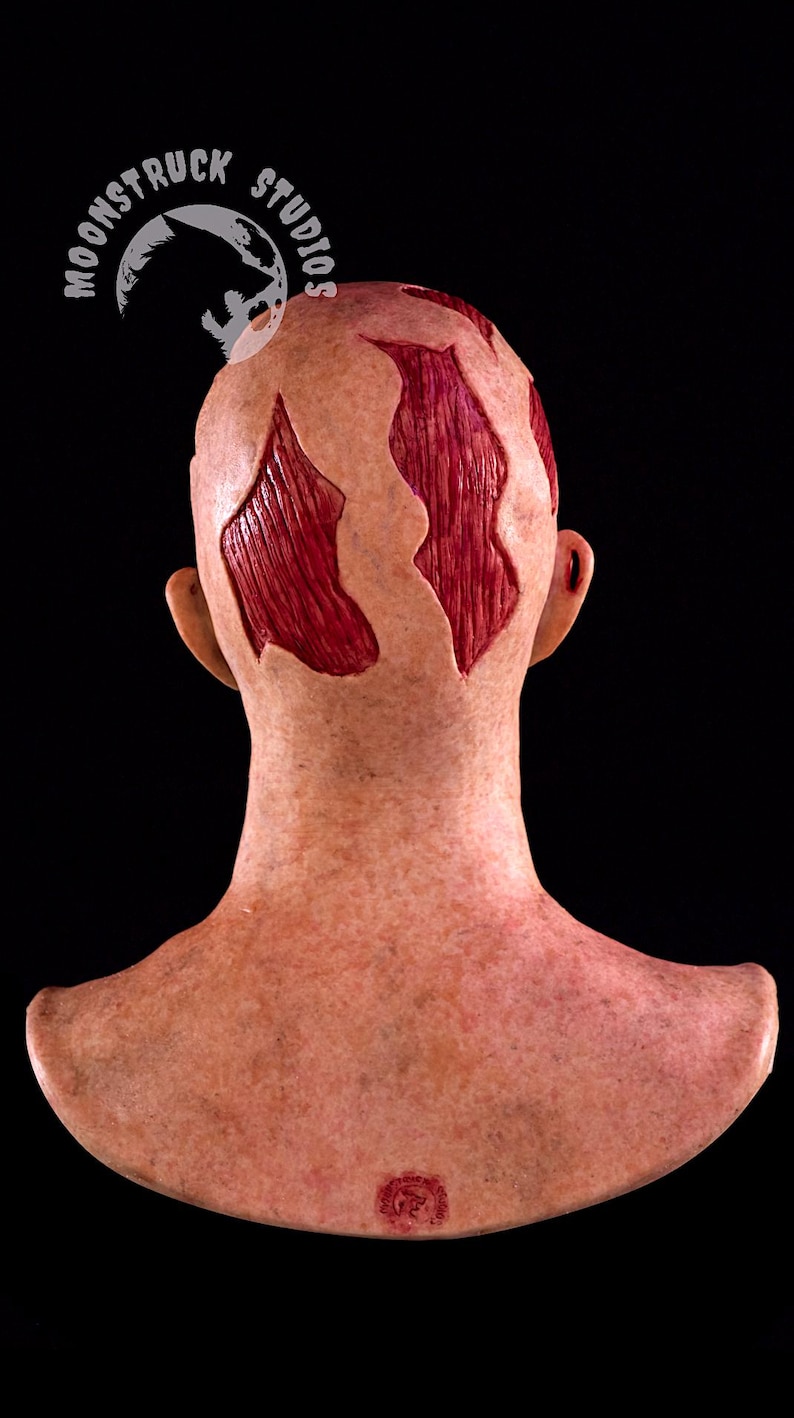 May include: A prosthetic head with exposed red muscle tissue, designed to simulate a scalp wound. The head is flesh-toned with areas of exposed muscle. The Moonstruck Studios logo is visible.