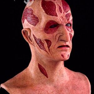 May include: A detailed mask of a character with a scarred face and exposed muscle tissue. The mask is flesh-toned with red accents, featuring deep cuts and wrinkles. The eyes are dark, and the overall effect is unsettling. The logo "MOONSTRUCK STUDIOS" is visible.