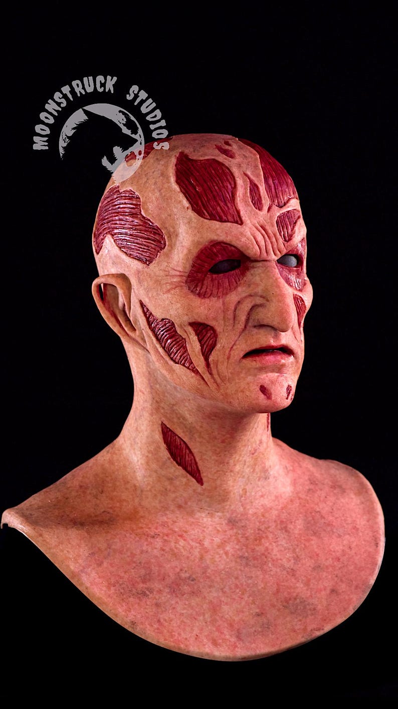 May include: A detailed mask of a character with a scarred face and exposed muscle tissue. The mask is flesh-toned with red accents, featuring deep cuts and wrinkles. The eyes are dark, and the overall effect is unsettling. The logo "MOONSTRUCK STUDIOS" is visible.