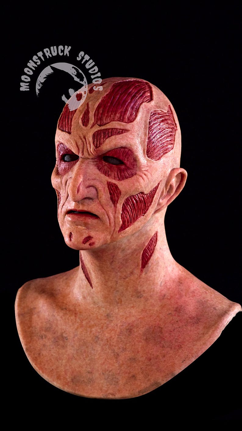 May include: A detailed, realistic mask depicting a scarred face with exposed muscle tissue. The mask features a pale complexion with deep red lacerations and exposed muscle. The eyes are dark and sunken. The Moonstruck Studios logo is visible.