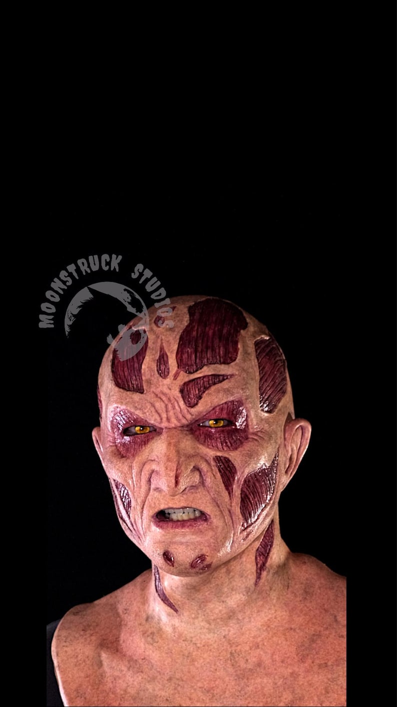 May include: A detailed mask depicting a scarred face with exposed muscle tissue. The mask features yellow eyes, a furrowed brow, and an open mouth, set against a black background. The logo "MOONSTRUCK STUDIOS" is visible.