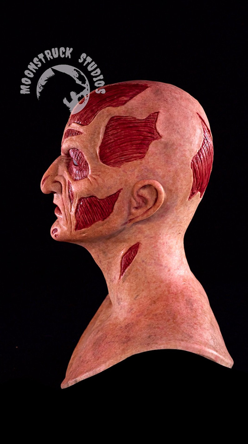 May include: A side profile of a gruesome mask with exposed muscle and skin. The mask is flesh-toned with red muscle details and a scarred appearance. The mask has a realistic, horror-themed design, perfect for Halloween or cosplay.
