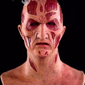 May include: A detailed, realistic mask with a scarred, weathered appearance. The mask features exposed muscle-like textures in shades of red and pink, with deep cuts and wrinkles across the face. The eyes are dark, and the overall effect is unsettling.