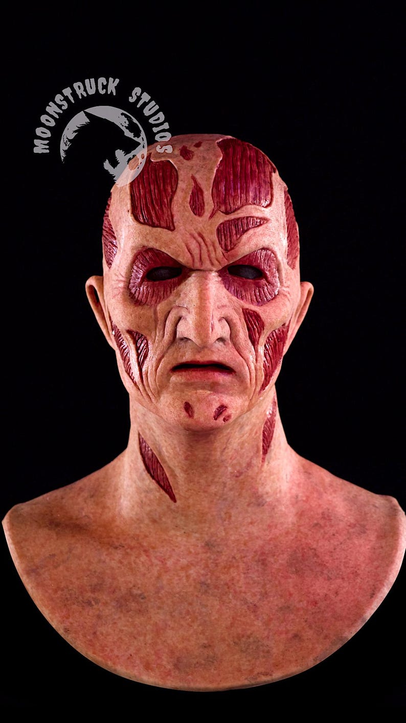 May include: A detailed, realistic mask with a scarred, weathered appearance. The mask features exposed muscle-like textures in shades of red and pink, with deep cuts and wrinkles across the face. The eyes are dark, and the overall effect is unsettling.