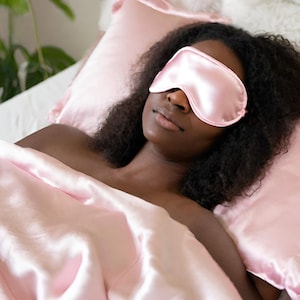 May include: A person sleeping in a bed with a pink satin sleep mask over their eyes. The bed is covered in a pink satin sheet and pillow.