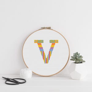 May include: A cross-stitch embroidery hoop displaying the letter "V" in a colourful rainbow design. The embroidery is set against a white backdrop, within a wooden hoop. A small succulent in a white geometric pot and scissors are also visible.
