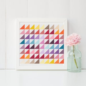 May include: A framed cross-stitch pattern with a geometric design of colorful triangles. The pattern is in a white frame and is sitting on a white surface.