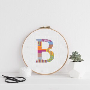 May include: A cross-stitch embroidery hoop displaying the letter "B" in a vibrant patchwork design. The letter is stitched with a variety of colours including blue, orange, pink, and yellow. A small succulent in a white pot and scissors are also present.