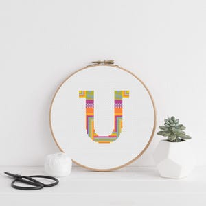 Puede incluir: Un bastidor de bordado de punto de cruz colorido con la letra "U". La letra está llena de un vibrante patrón de naranja, verde, morado y amarillo. También se ve una pequeña suculenta en una maceta geométrica blanca y unas tijeras.