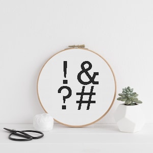 Peut inclure: Un cercle de broderie au point de croix avec des points de ponctuation noirs : point d'exclamation, esperluette, point d'interrogation et dièse. Une petite plante succulente dans un pot géométrique blanc est à droite. Ciseaux et fil au premier plan.