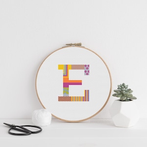 Peut inclure: Un cercle de broderie au point de croix avec la lettre "E" dans un motif patchwork multicolore. L'image présente également une petite plante succulente dans un pot géométrique blanc et une paire de ciseaux.