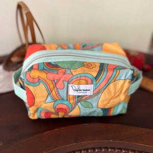 May include: A rectangular, zippered bag with a retro floral pattern in orange, yellow, and teal. The bag has teal handles and a teal zip. A small tag reads "Taylor Maker Handmade".