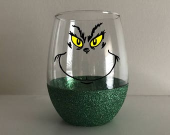 Op de afbeelding: Een groen glitter wijnglas met een zwart-wit Grinch-gezichtsontwerp. De Grinch heeft gele ogen.