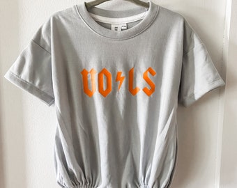 May include: A light gray baby onesie with short sleeves and a gathered bottom. The onesie has an orange graphic that says "VOLS" with a lightning bolt.