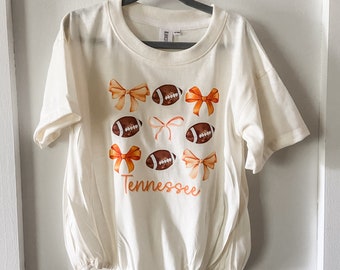May include: White baby onesie with orange and brown footballs and bows. The text "Tennessee" is printed on the onesie.